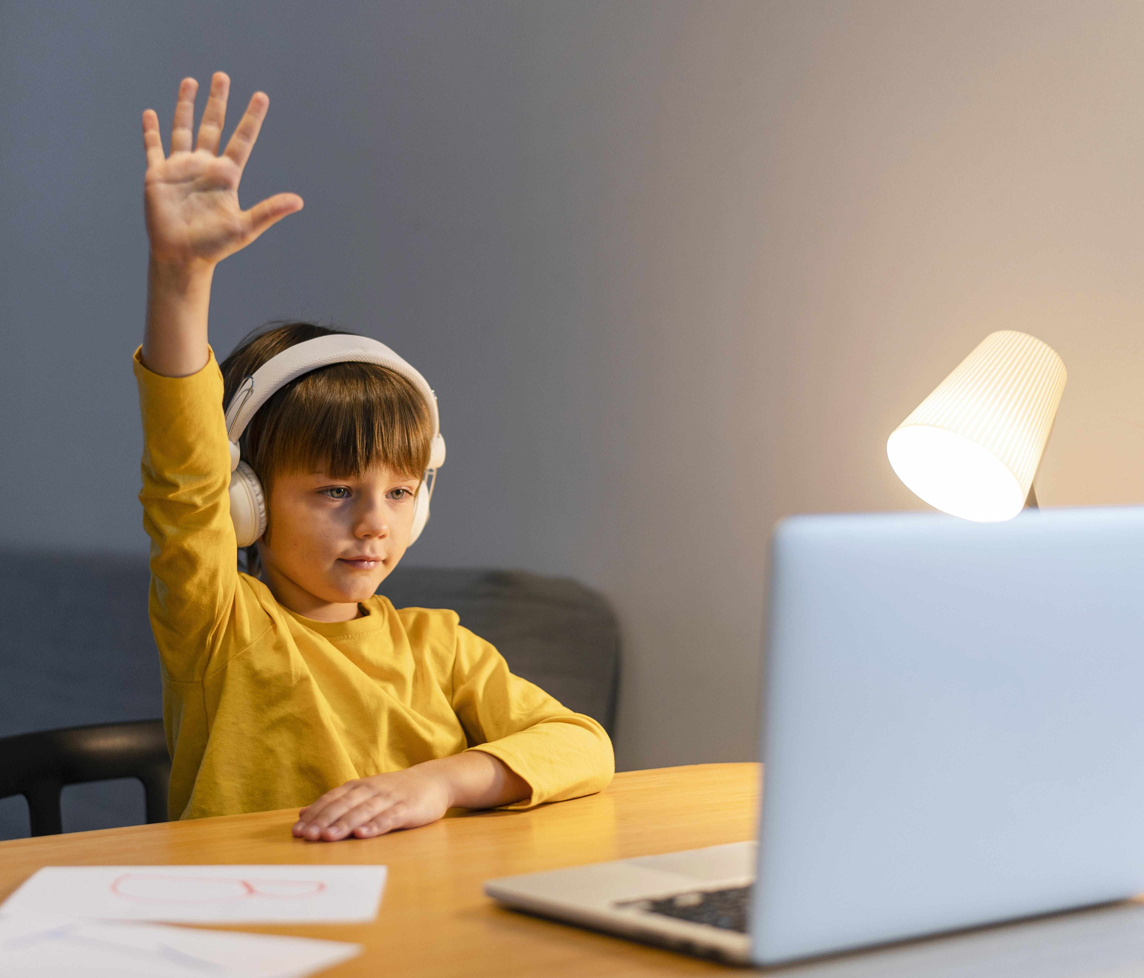 school boy yellow shirt taking virtual classes raising hand